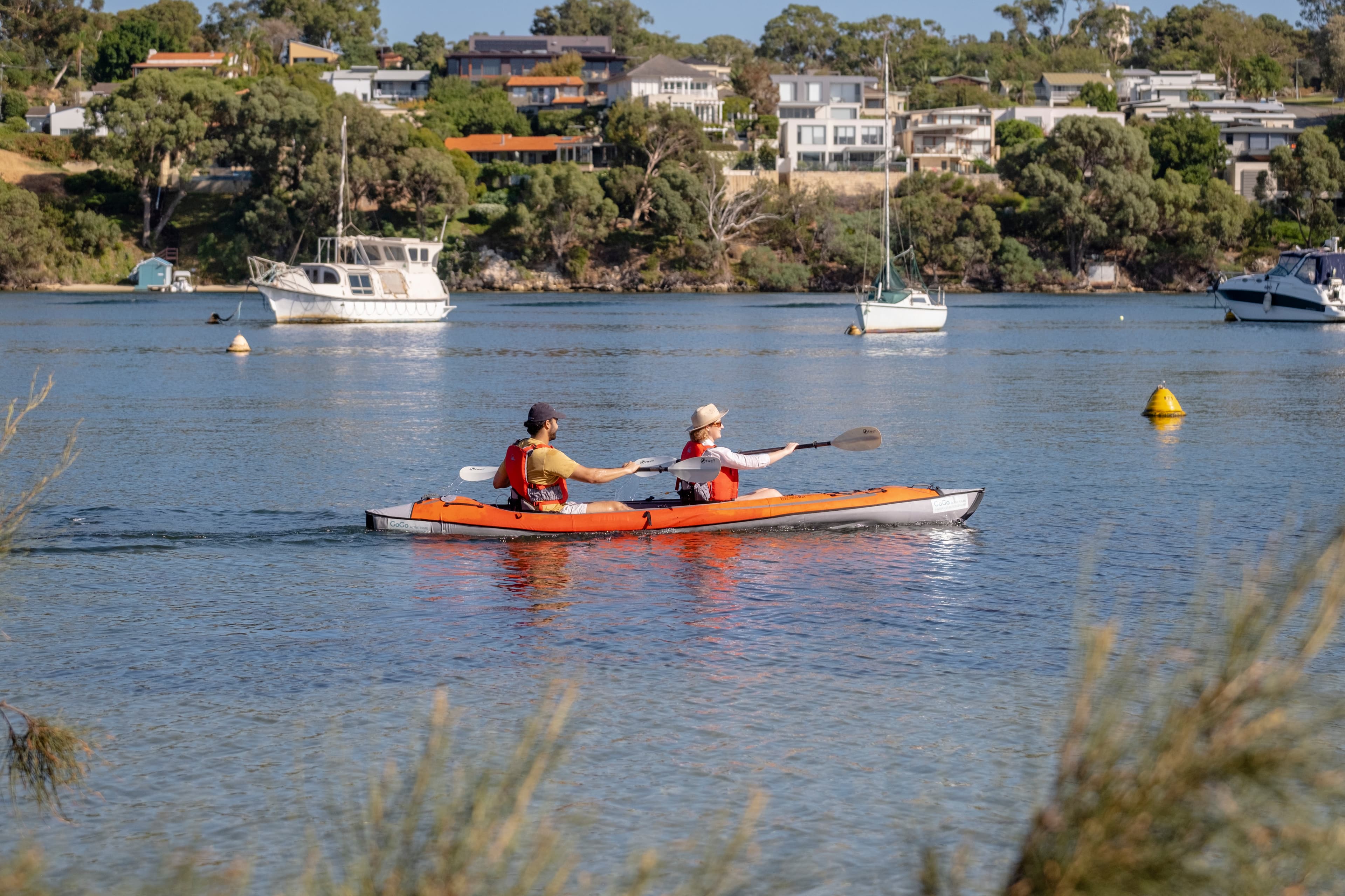 Two people kayaking in an orange kayak on a calm lake, with boats and houses on a tree-lined shore in the background.